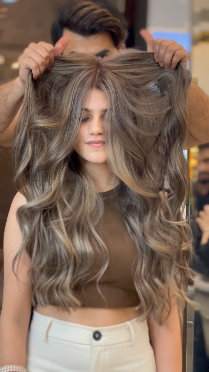 Long wavy ash-brown hair with blonde highlights lifted by a stylist in a salon