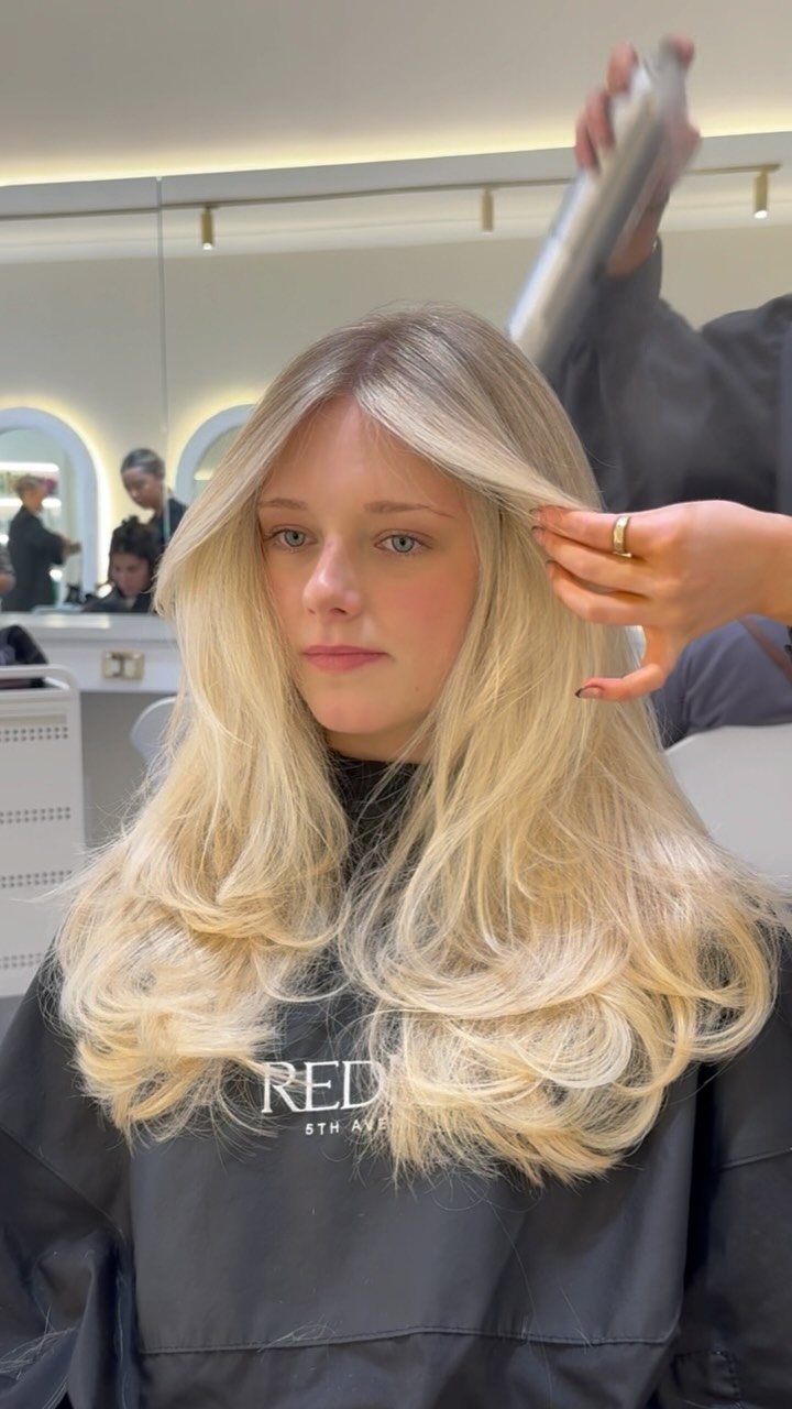 Woman with long, layered pale blonde hair seated in a salon chair