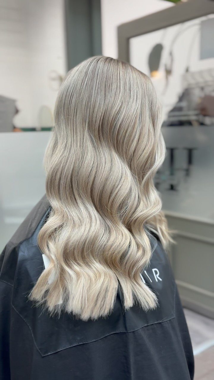 Long ash blonde waves with subtle fine highlights over a black salon cape
