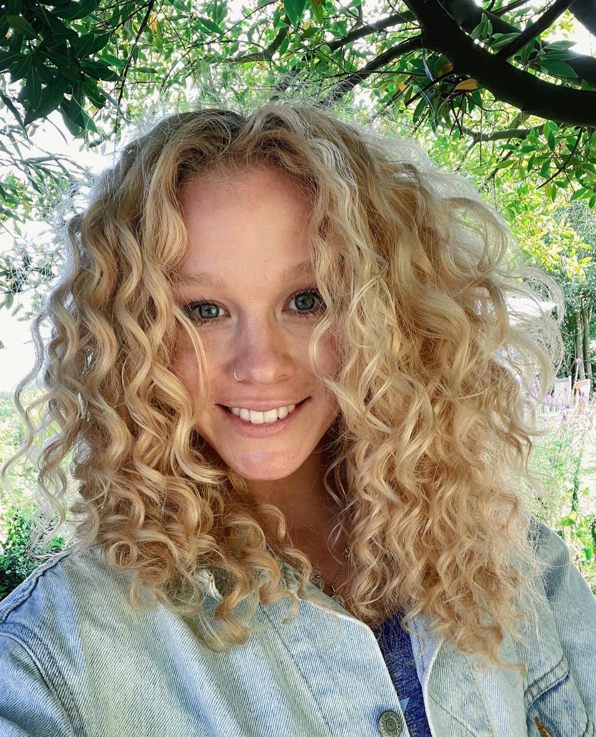 Woman with long blonde curls smiling beneath leafy branches outdoors