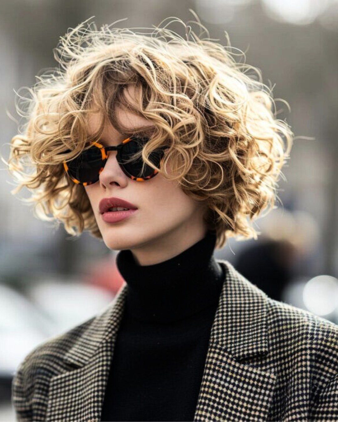 Short blonde curly bob with bangs frames a person wearing sunglasses and a checked coat.