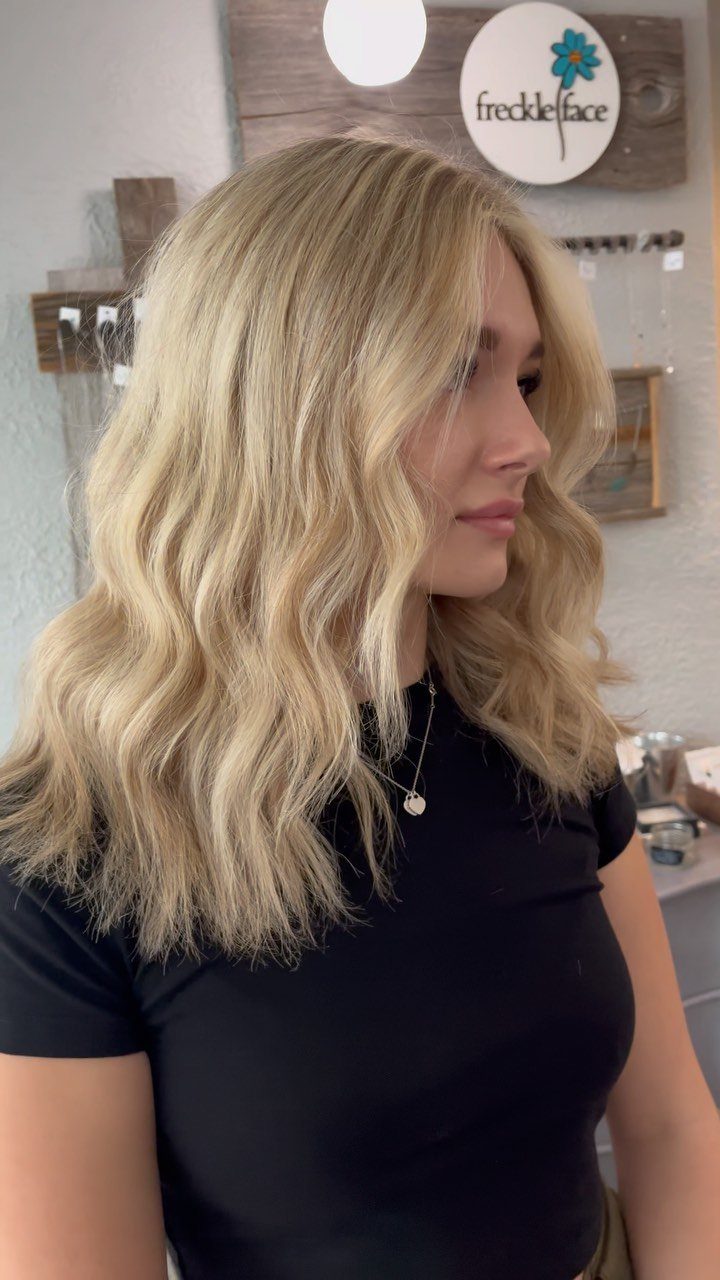 Wavy blonde hair with golden highlights falls over a black salon top