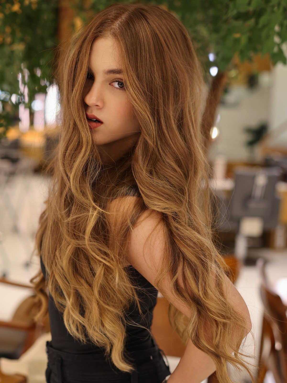 Woman posing with long, warm caramel blonde waves against a leafy café background