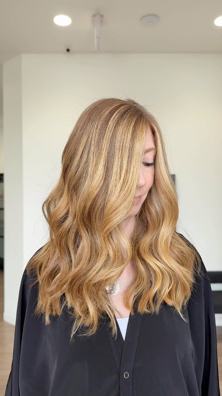 Long wavy golden blonde hair with warm coppery highlights shown against a black salon cape