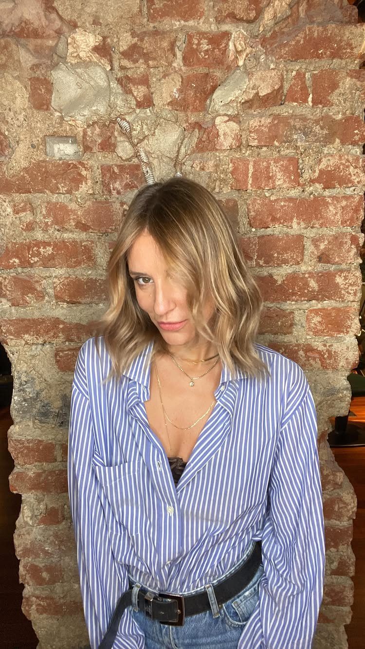 Woman with shoulder-length dirty blonde hair wearing a blue striped shirt against a brick wall