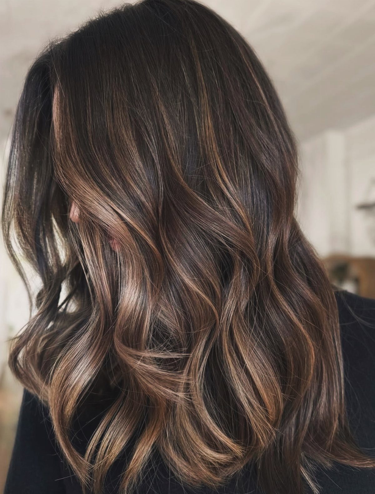 Long wavy dark hair with cool caramel highlights against a blurred indoor background