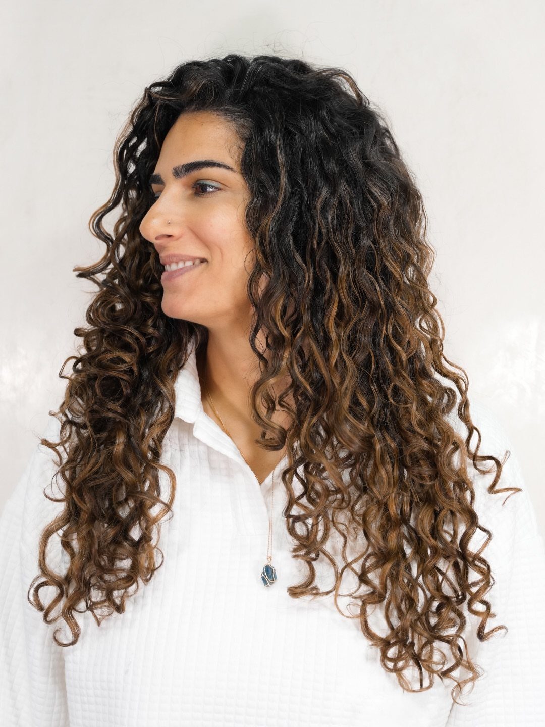 Long dark curls with warm caramel highlights frame a person in a white shirt