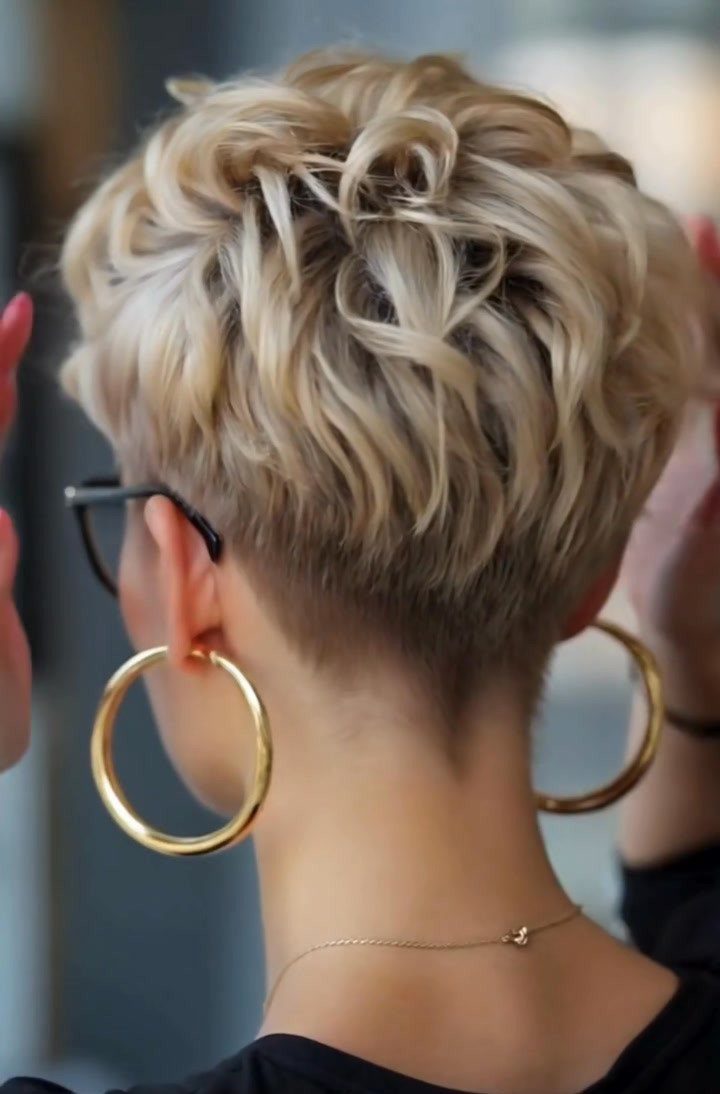 Short layered blonde curls with closely cropped sides, black glasses, and gold hoop earrings