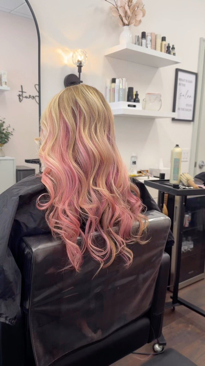 Long wavy blonde hair with pink ends drapes over a black salon cape