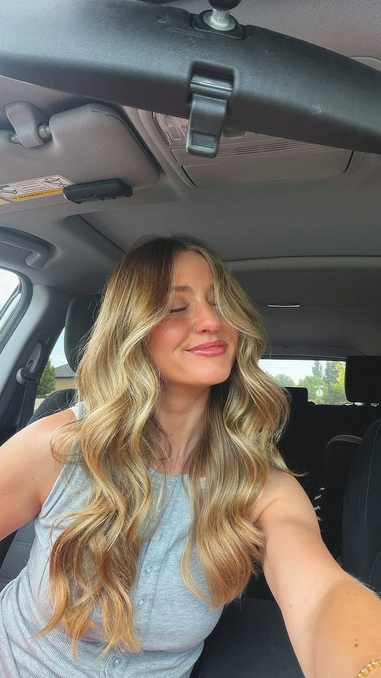 Woman with long wavy blonde hair and darker roots sitting in a car
