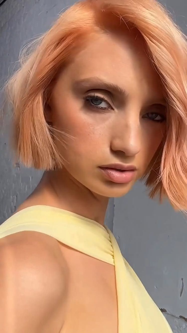 Woman with a chin-length peach-orange blonde bob poses against a gray wall