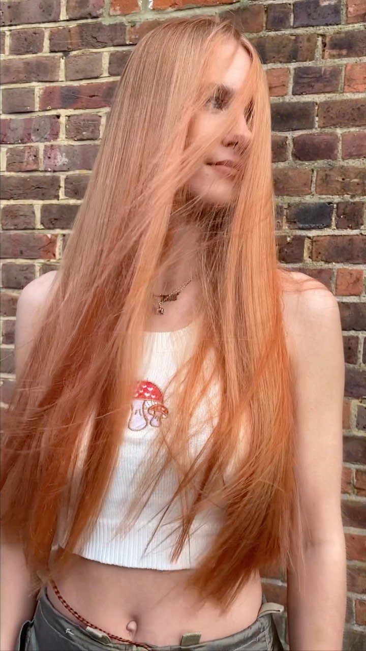 Woman with long, straight peach-copper blonde hair against a brick wall