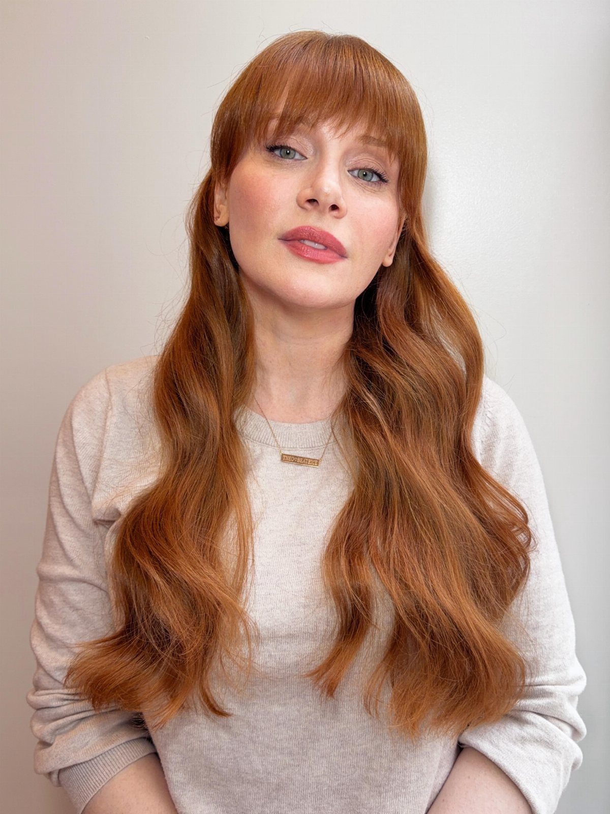 Woman with long, wavy strawberry-blonde hair and bangs wearing a pale sweater
