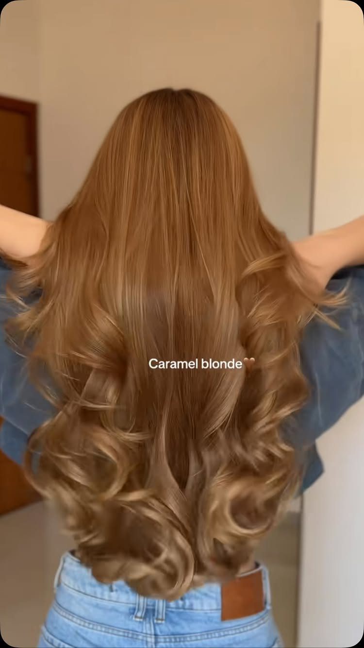 Long caramel blonde hair falls in glossy, loose curls down a person’s back