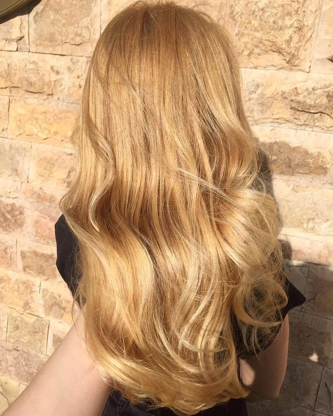 Long wavy golden blonde hair catches sunlight against a pale stone wall