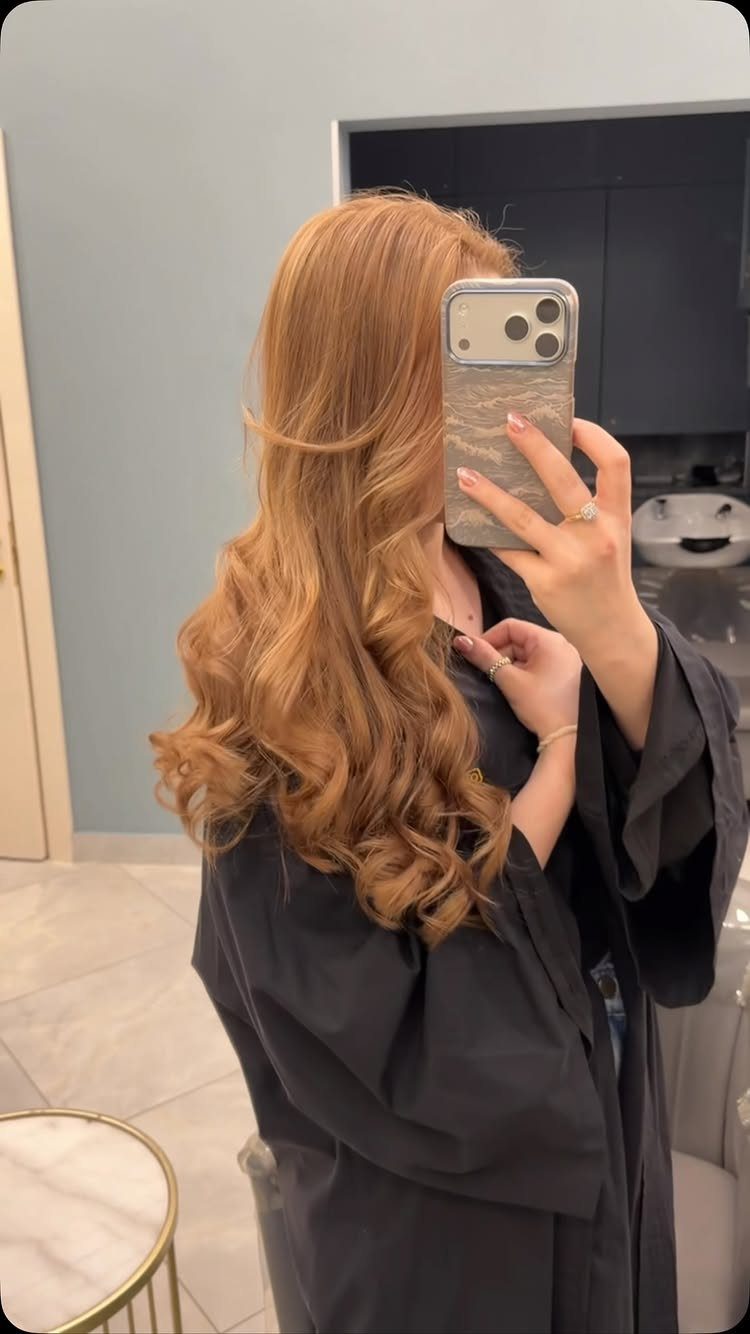 Long golden-blonde curls styled over a black salon cape in a mirror selfie