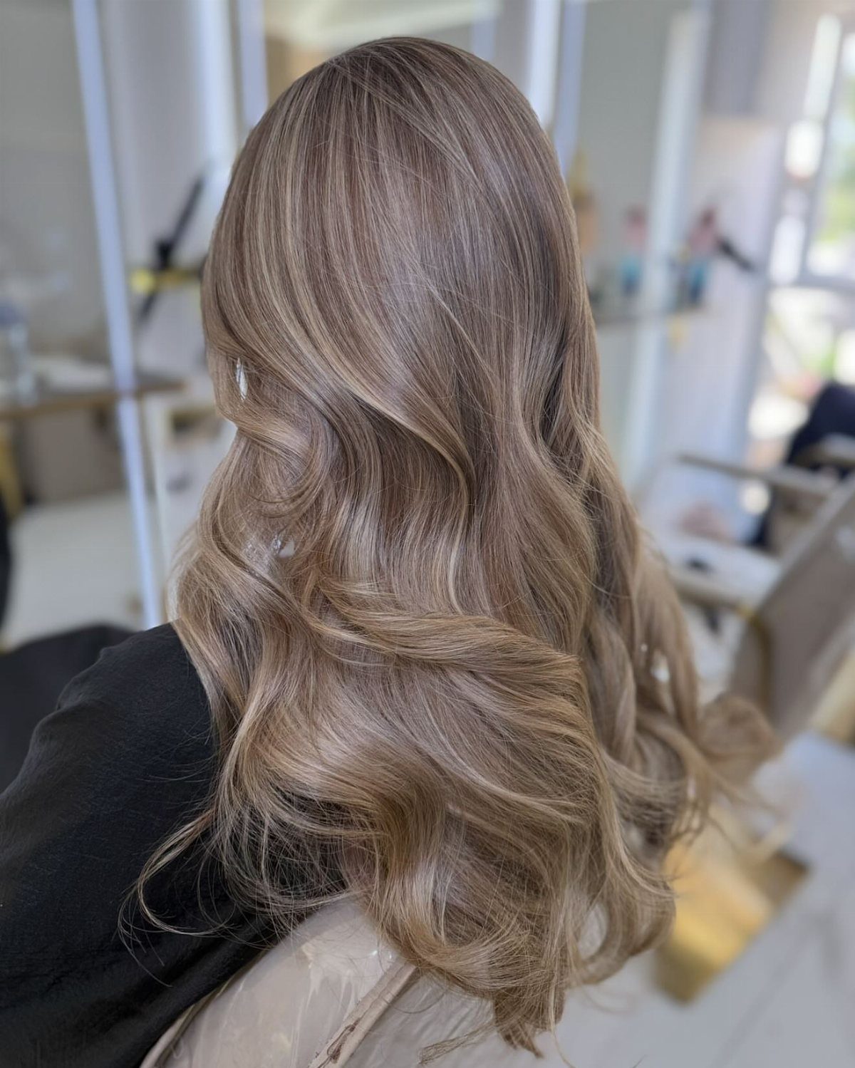 Long cool-toned brown and blonde balayage curls shown from behind in a salon