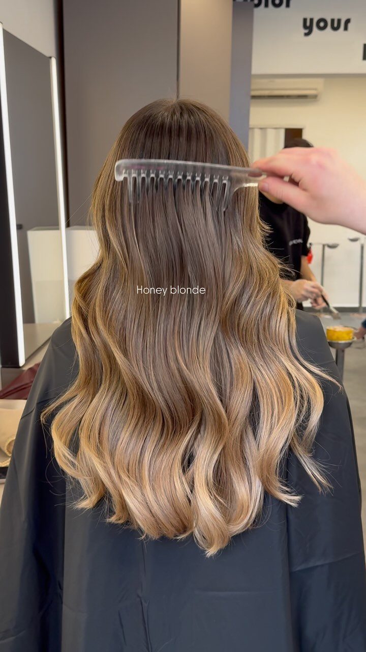 Long wavy hair fades from dark brown roots to honey blonde ends in a salon.