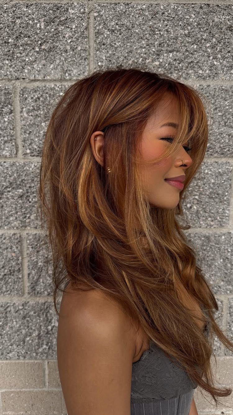 Woman with long layered honey-brown hair and side-swept bangs against a stone wall