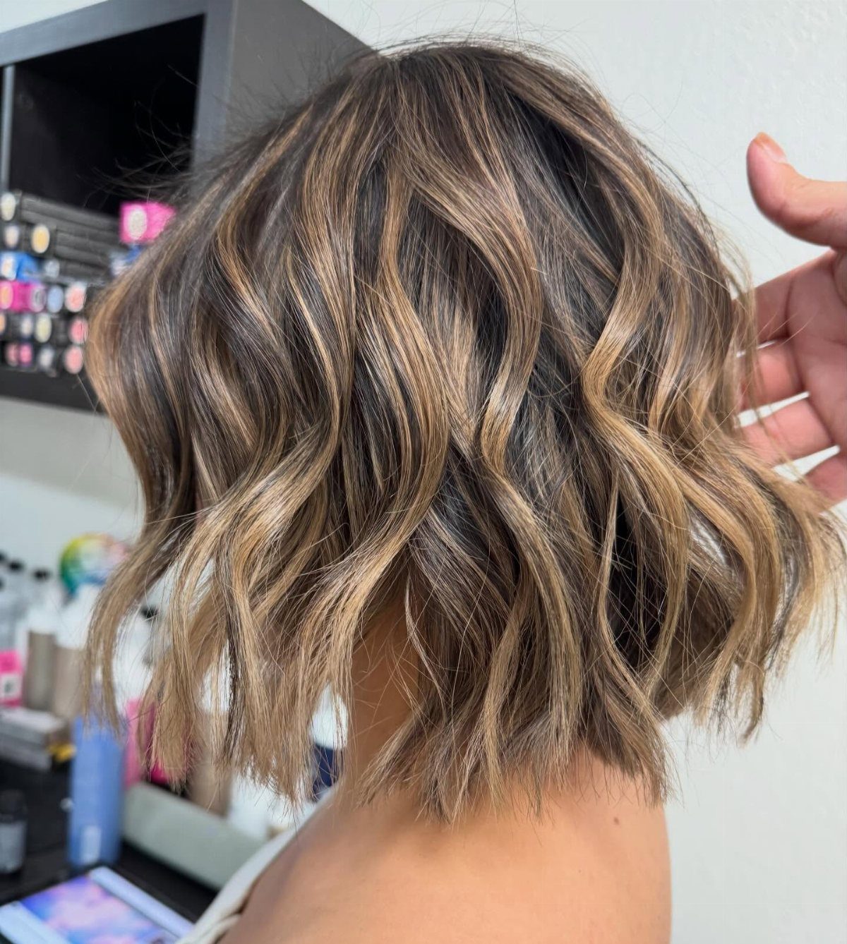 Wavy brunette bob with caramel honey ribbons throughout, lifted by a hand in a salon
