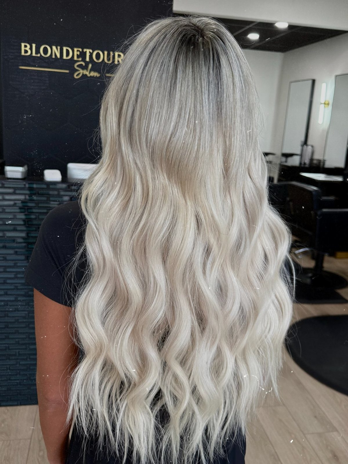 Long wavy blonde hair with dark roots displayed from behind in a salon