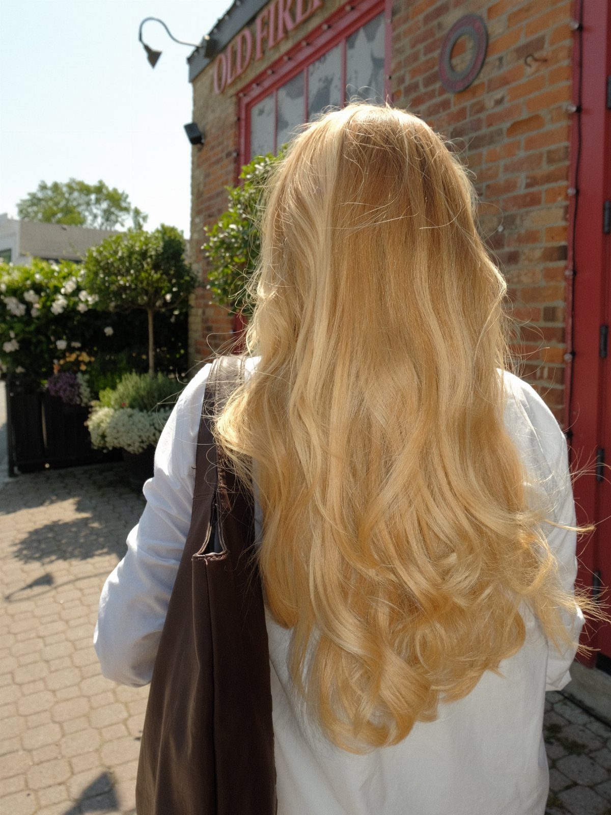Long golden-blonde waves fall over a white top beside a brick building
