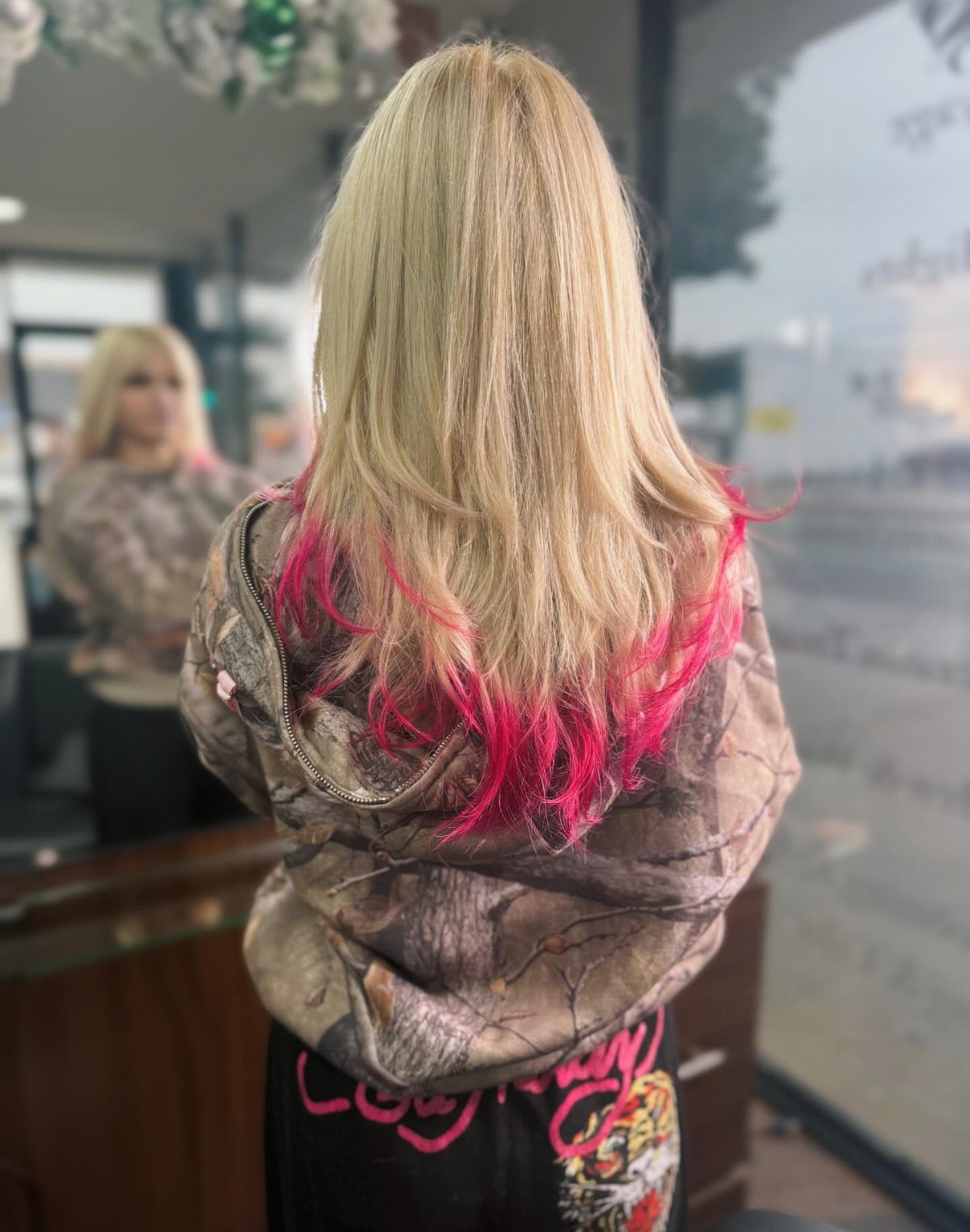 Long platinum blonde hair with vivid pink ends drapes over a camouflage jacket in a salon