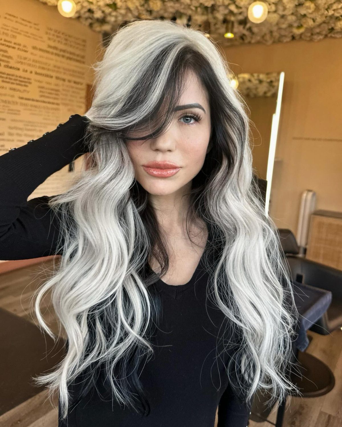 Woman displays long silver-white waves with prominent black roots in a salon