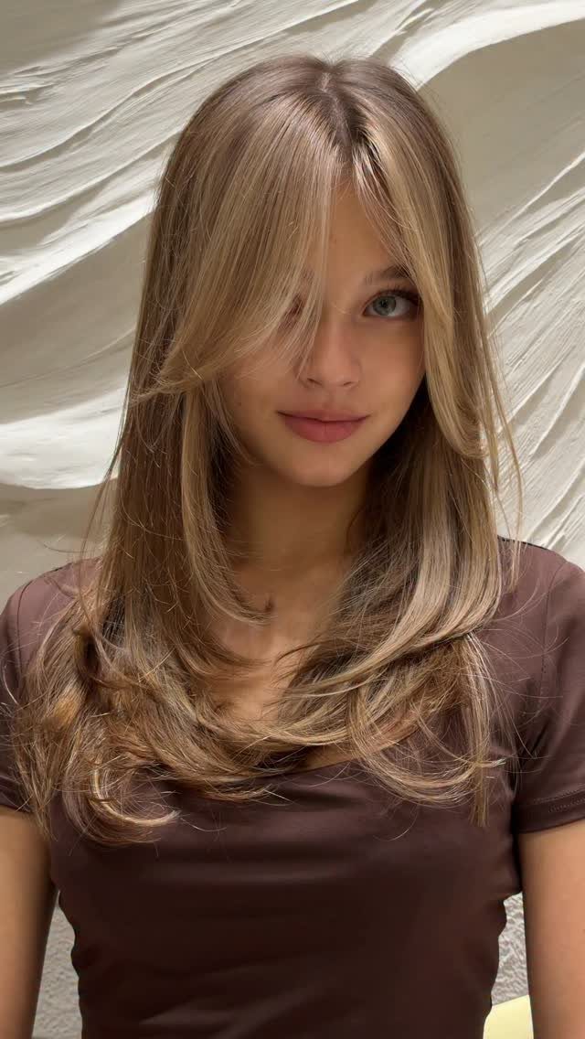 Woman with long, layered sand-blonde hair and curtain bangs against a light backdrop