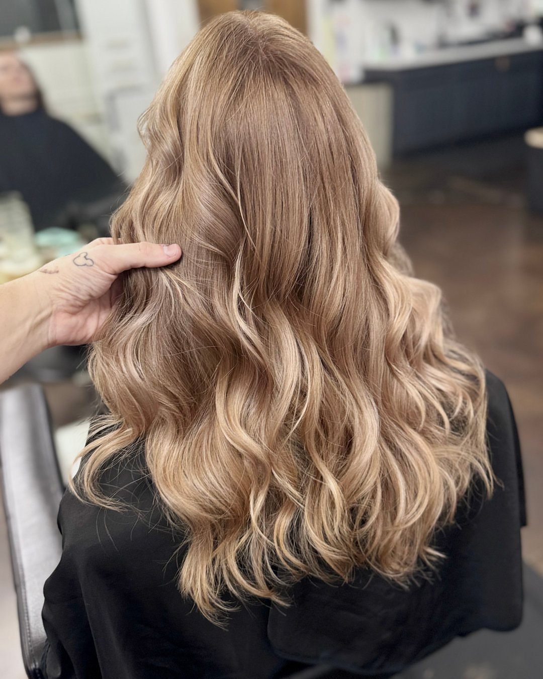 Long glossy sandy blonde waves drape over a salon chair as a hand touches the hair