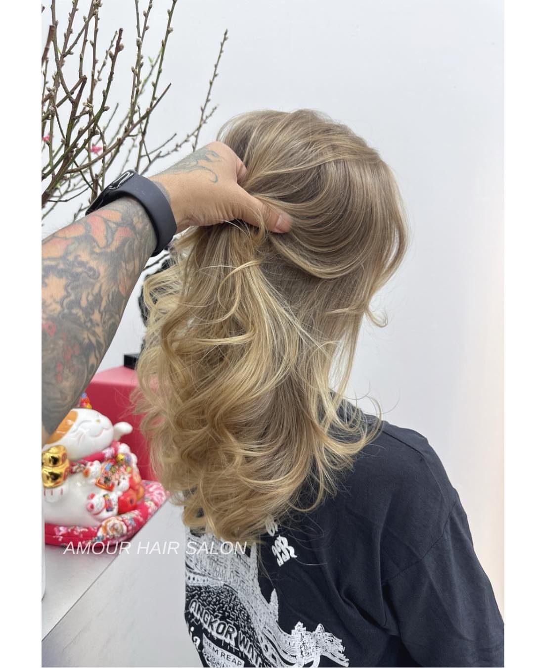 Long beige sandy blonde curls are lifted by a tattooed hand in a salon