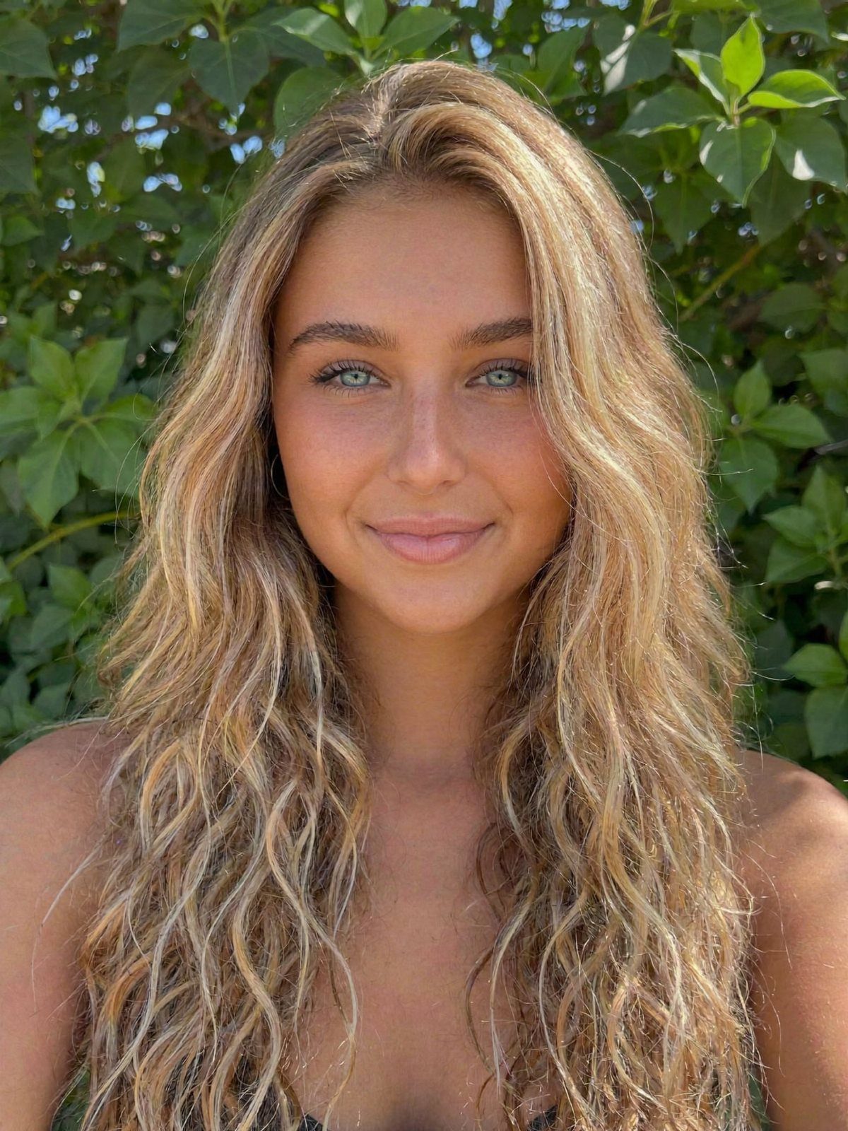 Woman with long, wavy golden sandy blonde hair posing against green foliage