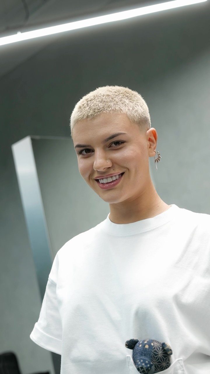 Person with closely cropped light blonde hair wearing a white T-shirt indoors