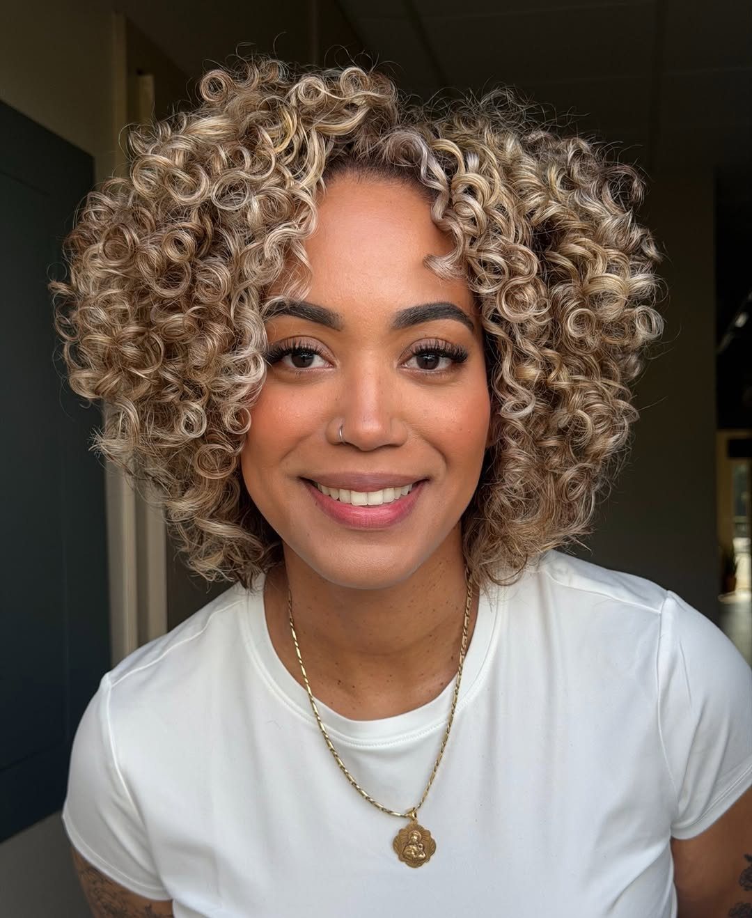 Woman with blonde face-framing curls wearing a white T-shirt and gold necklace